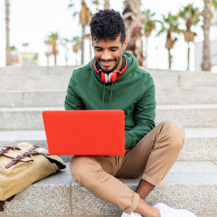 Portrait of hispanic young student working on laptop while sitting on steps outdoors - College, millennial people and education online concept