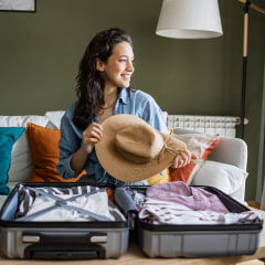 Young woman sitting on the couch and packing suitcase. She is holding a straw hat in her hands