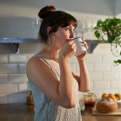 A woman standing in her kitchen drinks from a mug and the Keurig Cafe