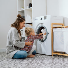 A caring mom with her little daughter sits in the bathroom, laundry at the washing machine, a sweet little girl sits on a woman's lap and puts towels in the open drum