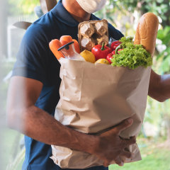 African American delivery man wearing protective face mask holding grocery paper bag of Healthy Food with fresh vegetables, milk, bread
