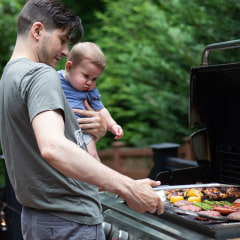 A man holding a baby, while grilling some meat and vegetables