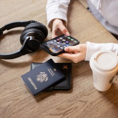 Woman at a coffee shop on her phone, surrounded by her passport and headphones