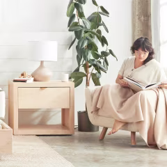 Illustration of a bed made with the Buffy Eucalyptus Duvet Cover, West Elm Cotton Linen & Velvet Corners Pillow Cover, Ekobo Recycled Bamboo Bambino Plate and a woman sitting in a chair next to her Avocado Malibu Wood Nightstand