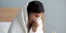 Close up sick woman wrapped into warm duvet blowing running nose, using paper napkin, holding cup of tea, sitting on bed at home, unhealthy young female feeling unwell, respiratory disease symptoms