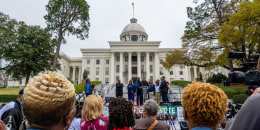 Civil rights activists speak at a podium to a crowd in front of the Alabama State Capitol