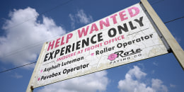 Large sign photographed from below against a blue sky with puffy clouds reads: Help Wanted Experience Only - Asphalt Luteman, Roller Operator, Pavebox Operator