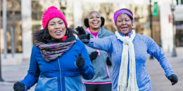 Three African-American women exercising together in the city, jogging or power walking, smiling. The one on the right, a mature woman in her 40s, is trying to catch up to her friend, a senior woman in her 60s, reaching to tap her on the shoulder.