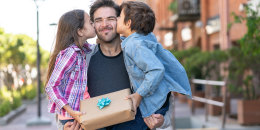 Loving son and daughter kissing daddy on the cheek while he carries them both with a present for father's day.