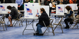 Voters fill out their ballots at a polling location behind dividers that say "Vote" with the American flag