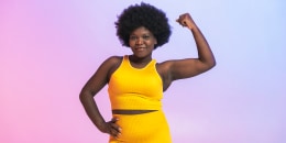 Smiling young woman in yellow sports clothing flexing muscles against two tone colored background.