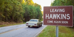 A car on a road passes by a red sign with white text that reads "Leaving Hawkins Come Again Soon"