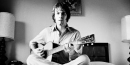 Bob Weir sits cross legged on a bed while strumming a guitar