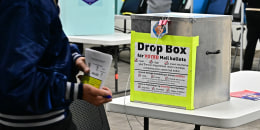 A man holding a voting ballot passes in front of a metal lockbox with a label reading DROP BOX FOR VOTED MAIL BALLOTS inside a polling station.