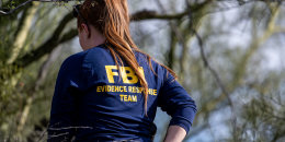 A woman with a long ponytail and a blue longsleeved shirt that says FBI EVIDENCE RESPONSE TEAM walks among trees.