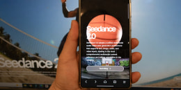 A hand holds up a mobile phone in front of a computer screen. Both read "Seedance 2.0".