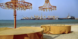 Two tables with chandeliers on a beach in front of large container ships floating in the distance.