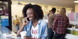 Image: Smiling young woman drinking coffee and talking on cell phone at laptop in cafe