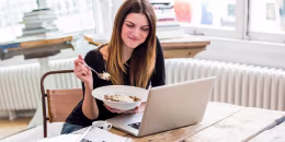 Young woman in city apartment eating muesli breakfast whilst reading laptop