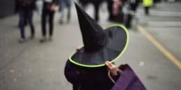 Child Holding onto Witch's Hat and Purple Bag on Urban Sidewalk, Rear View