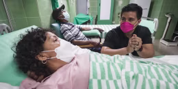 George Solis, right, wearing a pink mask, speaks with Yonelkys García who is laying in a hospital bed and wearing a white mask