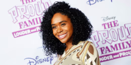 Raquel Lee Bolleau smiles while standing on the red carpet, the white backdrop behind her has text that reads "The Proud Family, Louder and Prouder" and "Disney" in pink and purple text