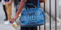 A guest wears a thin gold bracelet on the wrist, a large rectangular washed blue denim Telfar tote with dual top handles, a long shoulder strap, holding a clear plastic cup of iced coffee with a turquoise straw.