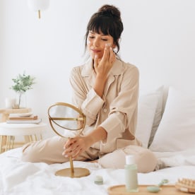 Woman doing self care applying beauty cosmetic, looking into mirror, while sitting in her bed