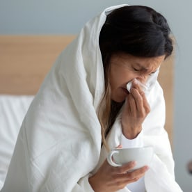 Close up sick woman wrapped into warm duvet blowing running nose, using paper napkin, holding cup of tea, sitting on bed at home, unhealthy young female feeling unwell, respiratory disease symptoms