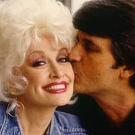Dolly parton smiles as a dark haired main kisses her cheek.