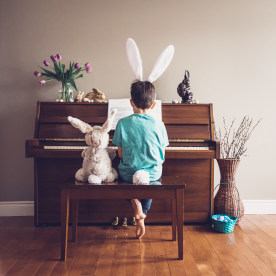 Boy wearing bunny ears playing piano with stuffed rabbit beside him.