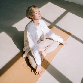woman with short blond hair is meditating sitting in lotus position on yoga mat in front of a window. 