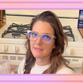 Drew Barrymore in glasses kneels on her kitchen floor in front of a white gas stove.