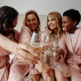 Women celebrate a bachelorette party of bride. Group of female sitting on bed and toasting champagne glasses at home. Focus on champagne flutes.