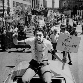 Harvey Milk sitting on top of a car during the Pride Parade. He is holding a placard that reads: 'I'm from Woodmere, NY'. 