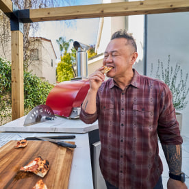 Jet Tila in a flannel shirt bites into a pizza in a backyard outdoor kitchen.