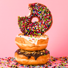 Three donuts with sprinkles on pile, one with missing bite, over pink background.