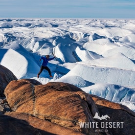 A man jumps on a rock in Antarctica in a blue puffer jacket. Behind him are white glaciers of snow.