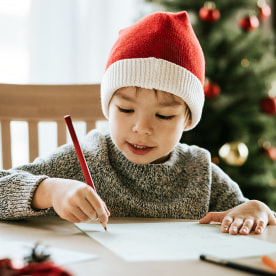 Boy writes a letter to Santa Clause with Santa hat and Christmas tree in the background.