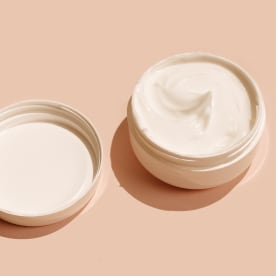 Face moisturizer in a white jar on a beige background.