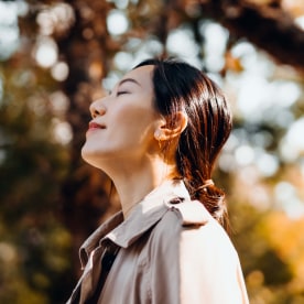 Portrait of young Asian woman having a walk in the park, enjoying the warmth of sunlight on a beautiful Autumn day outdoors and breathing fresh air with eyes closed. 