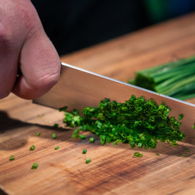 A person is cutting green herbs on a wooden cutting board with a knife.