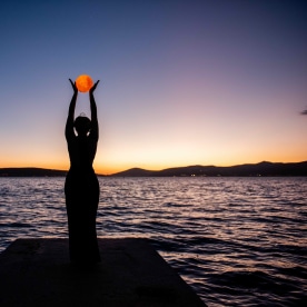 Silhouette of young woman in black dress, backdrop of twilight by sea, holding full moon in hands.