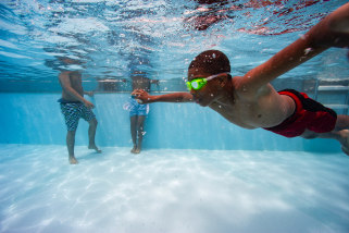 A child wearing goggles swims underwater in a pool, two other children are seen standing in the pool