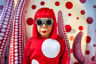 Yayoi Kusama stands in front of her artork, inflated red tubes with white polkadots, she has a bright red bob and sunglasses while wearing a bright read shirt with white polkadots