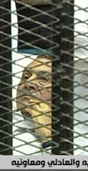Image: Hosni Mubarak laying on a hospital bed inside a cage of mesh and iron bars in a Cairo courtroom