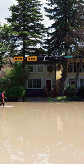 Image: A woman walks down a flooded street in the Elbow Park area of Calgary