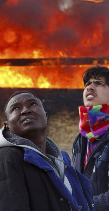 Image: Migrants stand next to a burning makeshift shelter during the dismantlement of the migrant shanty town called the \"Jungle\" in Calais