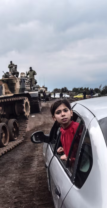 Image: A girl leans out of the window of a car to look as Turkish army tanks and soldiers gather close to the Syrian border