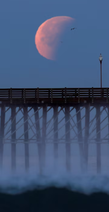Image: A Blue Moon comes out of a lunar eclipse as it sets past an ocean pier in Oceanside, California